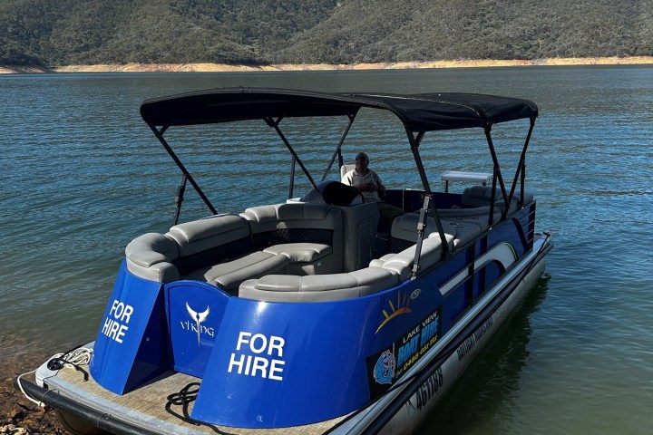 Blue pontoon boat labeled 'For Hire' docked by a lake with wooded hills in the background.