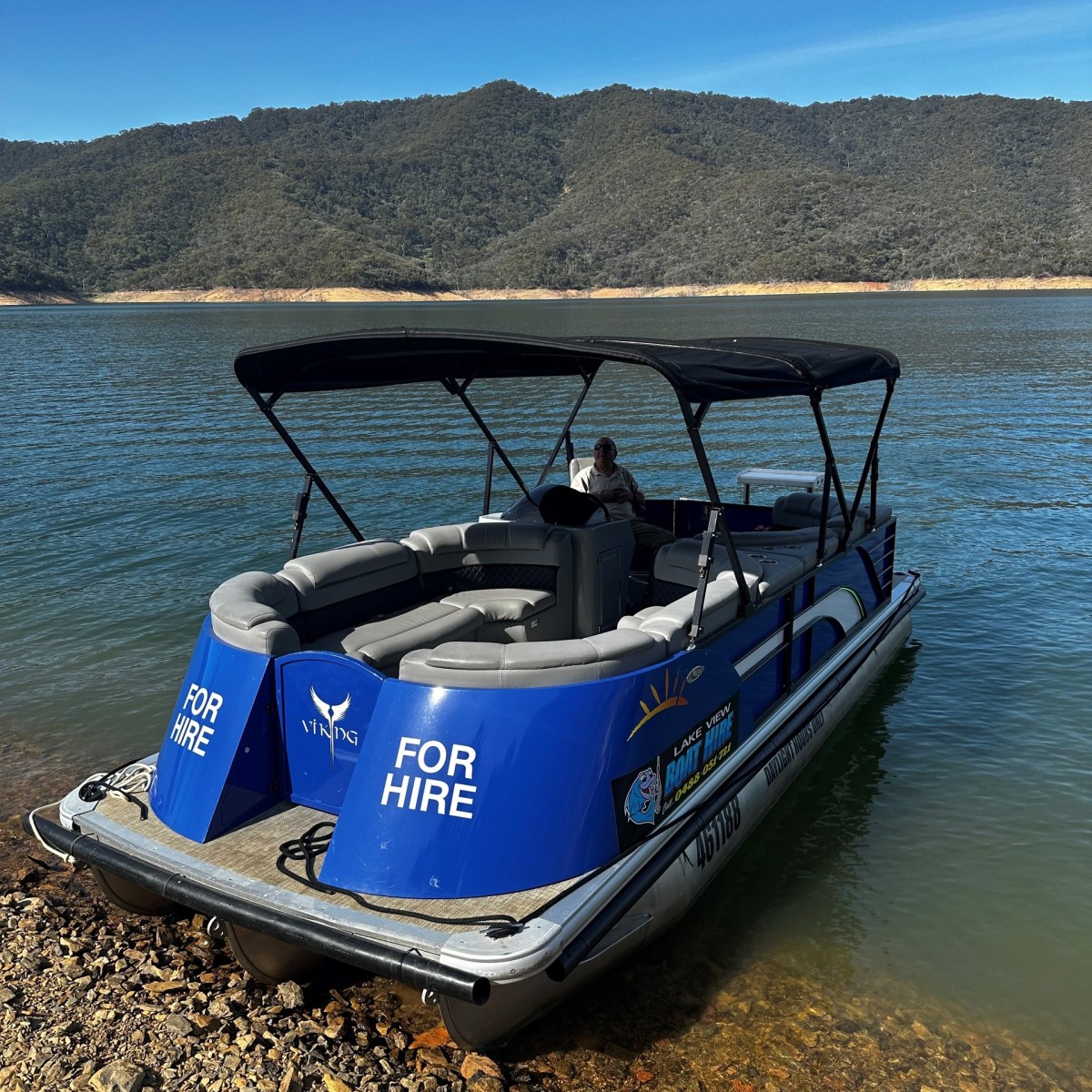 Blue pontoon boat labeled 'For Hire' docked by a lake with wooded hills in the background.