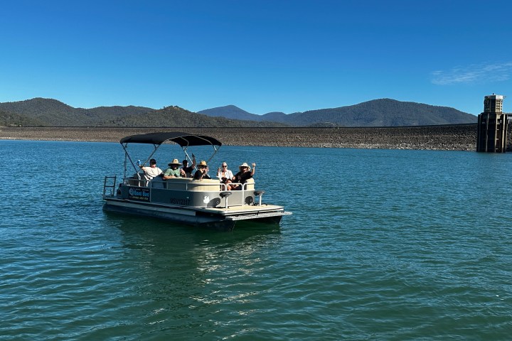 Group on pontoon boat in lake with mountainous background and clear blue sky.