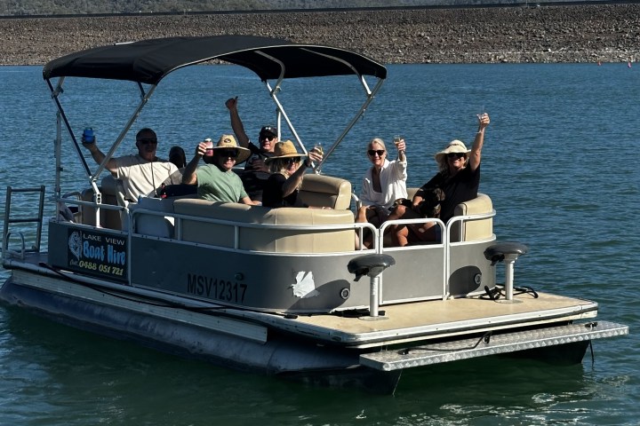 Group of people waving on a pontoon boat in a lake with mountains in the background.