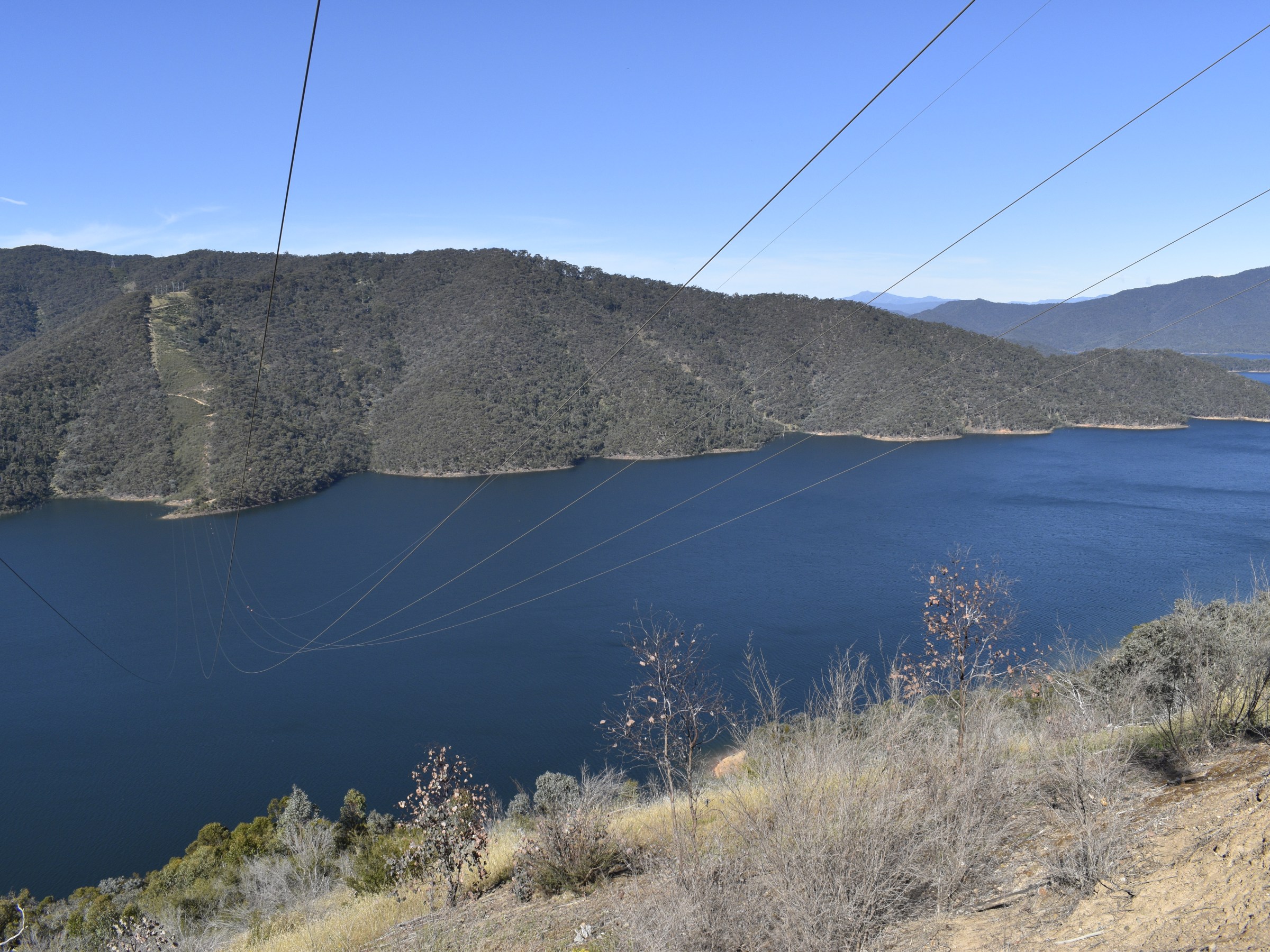 Overhead power lines above a lake with surrounding hills and clear blue sky.