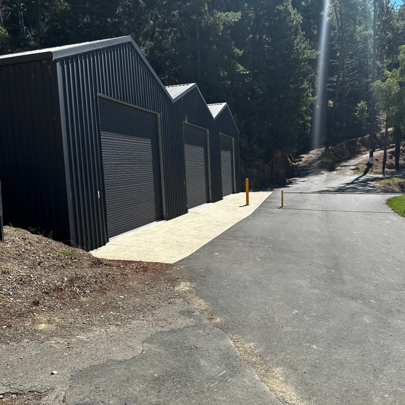 Three black garages with closed doors on a sunny day, trees in the background.